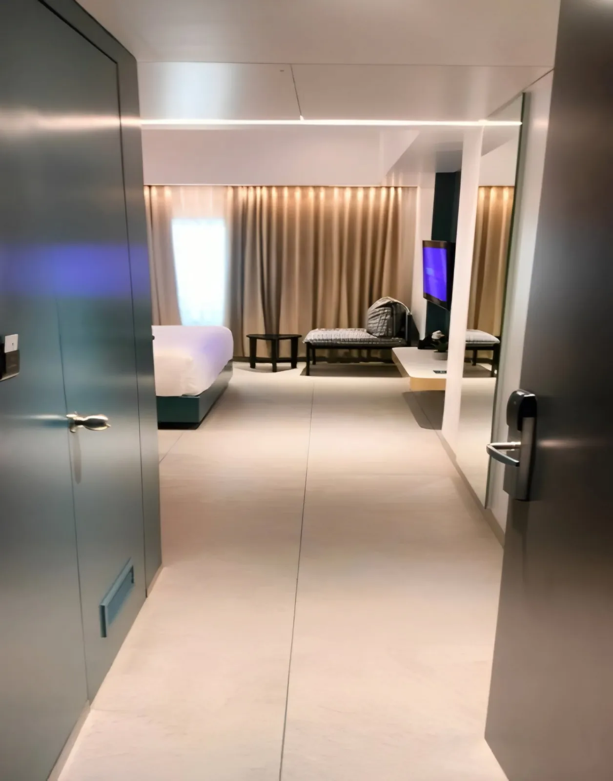 View from a guest room doorway across pale floor tiles to the bed and curtained window