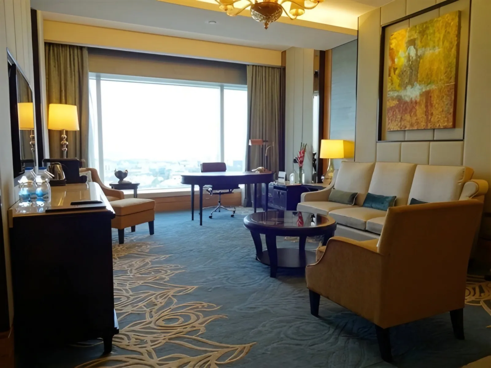 Hotel suite with sofas and armchair, a desk by a large window and a patterned blue carpet