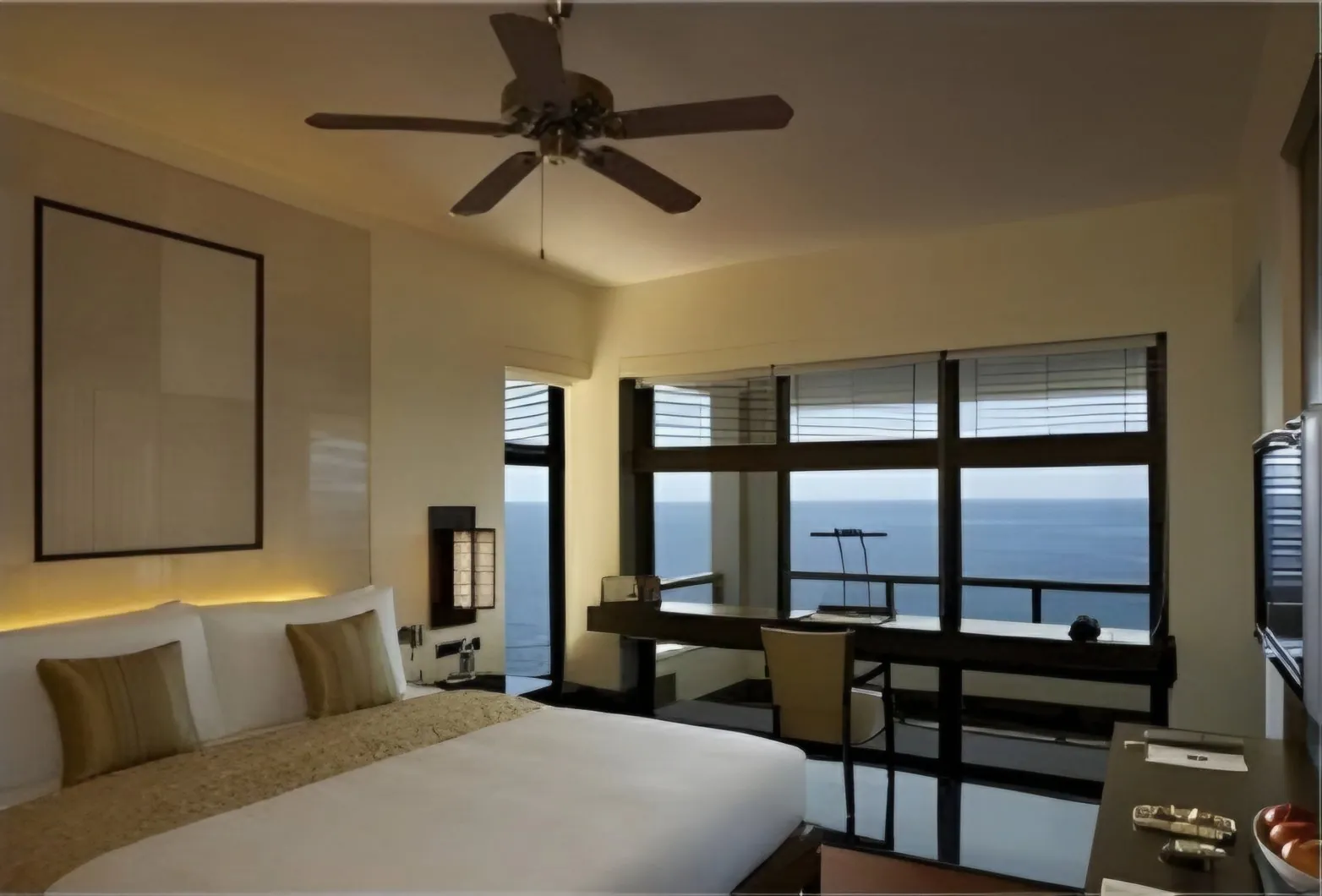 Resort bedroom with a ceiling fan and a desk facing floor-to-ceiling windows onto the sea